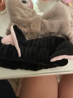 Två fluffiga hårband med rosett - Säljer två mjuka och fluffiga hårband i plysch, ett i beige med stor rosett och ett svart med rosa insida och kattöron. Perfekta för att hålla håret borta vid sminkning eller hudvård. Supersöta accessoarer som ger en gullig touch till din look.