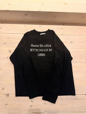Svart långärmad topp Acne Studios - Svart långärmad topp från Acne Studios med tryck i vitt framtill där det står 'Acne Studios STOCKHOLM 1996'. Rund halsringning och avslappnad passform. Perfekt för en stilren och trendig look.