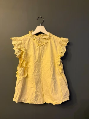 Gul volangblus från H&M - Söt gul blus från H&M med volangdetaljer och broderade spetsar längs ärmhålen. 