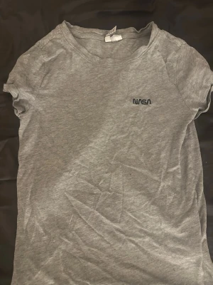 Grå NASA t-shirt med tryck - Säljer en grå t-shirt med svart NASA-tryck på bröstet. T-shirten har rund hals och korta ärmar. Tillverkad i mjukt bomullsmaterial och har en enkel, stilren design som passar perfekt till jeans eller shorts.