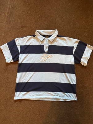 Ed hardy x Urban outfitters Shirt - Säljer en kortärmad pikéskjorta med breda marinblå och vita ränder. Skjortan har krage, knappar framtill och ett broderat motiv på bröstet samt en stor broderad ros på ryggen. Perfekt för en avslappnad och stilren look. Aldrig använt! Pris kan diskuteras