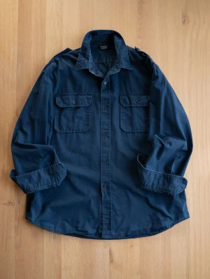 Mörkblå overshirt med fickor - Stilren mörkblå overshirt i bomull med två bröstfickor med lock och knappar. Klassisk krage och knäppning framtill. Perfekt lager-på-lager-plagg för en avslappnad look. Ärmarna kan vikas upp för en mer casual stil.