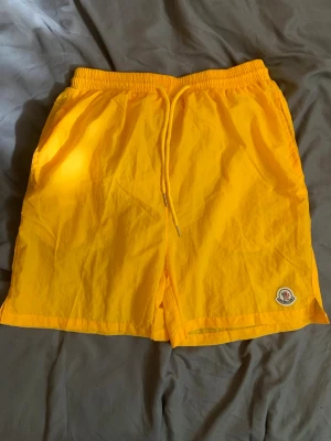 Gula badbyxor från Moncler - Snygga gula badbyxor från Moncler med elastisk midja och dragsko. Klassisk Moncler-logga på vänster ben. Tillverkade i lätt och snabbtorkande material, perfekta för stranden eller poolen.