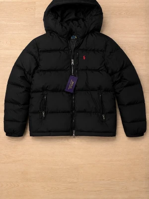 Svart pufferjacka från Polo Ralph Lauren - Snygg svart pufferjacka från Polo Ralph Lauren med huva och två dragkedjeförsedda fickor. Jackan har en röd logga broderad på bröstet och quiltad design som ger en klassisk look. Perfekt för kalla vinterdagar och riktigt varm.