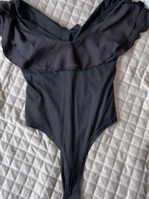Svart body med volang  - Stilren svart body med bred volang längs axlarna och v-ringad hals. Modellen är åtsittande med hög benskärning och ger en elegant siluett. Tillverkad i mjukt stretchigt material för skön passform. Precis som den i vit men svart. 