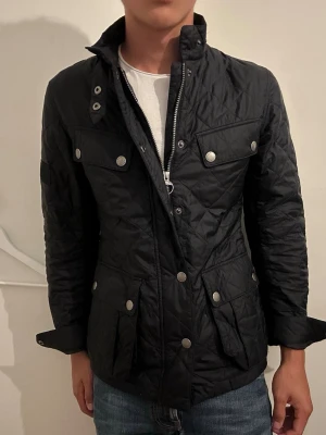 Svart quiltad jacka med fickor - Snygg svart quiltad jacka med hög krage och tryckknappar. Fyra framfickor med lock och markerad midja ger en schysst siluett. Jackan har dragkedja och knappar framtill samt detaljer på ärmen. Perfekt för dig som vill ha en stilren och funktionell jacka.