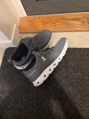 Gråa On Cloud sneakers - Gråa On Cloud sneakers med vit sula och ventilerande mesh-ovandel. Skorna har snörning, diskret logga på sidan och en unik, dämpad sula med tydliga rutor. Perfekta för dig som gillar sportig och stilren look.