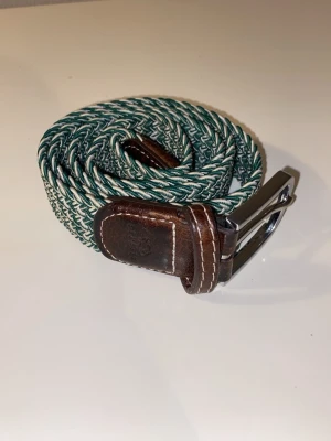 Grönflätat bälte från Billybelt - Snyggt flätat bälte från Billybelt i grönt och vitt med detaljer i brunt skinn. Bältet har ett klassiskt spänne i silverfärgad metall och en flexibel, vävd design som gör det enkelt att justera. Jätte bra kvalite! Säg till om ni undrar något🙌