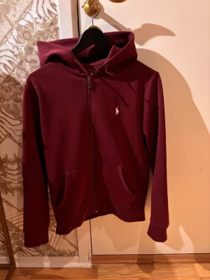 Vinröd hoodie med dragkedja Polo Ralph Lauren - Snygg vinröd hoodie från Polo Ralph Lauren med dragkedja framtill och två fickor. Klassisk logga broderad på bröstet. Perfekt för en avslappnad och stilren look.