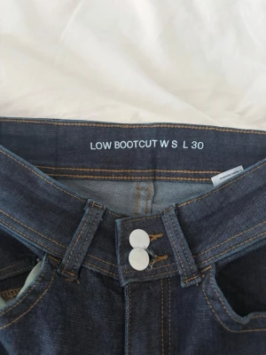 Mörkblå bootcut jeans low waist - Säljer ett par mörkblå bootcut jeans med låg midja och raka ben. Jeansen har dubbla silverknappar i midjan och klassiska fickor fram och bak. Snygga kontrastsömmar och tillverkade i denim. Perfekta för en avslappnad och trendig look.