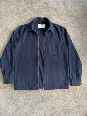 Mörkblå overshirt från A Day's March - Stilren mörkblå overshirt från A Day's March i mjukt tyg. Klassisk krage, knappstängning vid ärmslut och en bröstficka. Perfekt lager-på-lager-plagg med enkel design och vanlig passform.