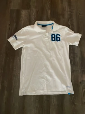 Vit pikétröja med blå detalj och 86 - Säljer en vit pikétröja med blå detaljer och siffran 86 broderad på bröstet. Tröjan har klassisk krage, korta ärmar och knappslå framtill. Perfekt för en sportig och avslappnad stil.