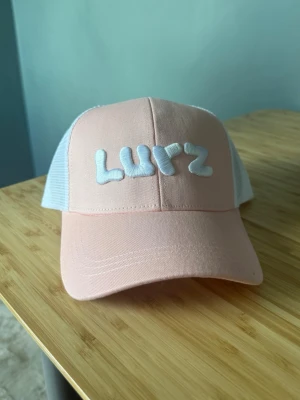 Lurz keps - Snygg rosa och vit truckerkeps med broderad texten 'Lurz' framtill. Kepsen har vit mesh baktill för extra ventilation och justerbar passform. Perfekt accessoar för en avslappnad streetwear-look. Helt ny, passade inte min huvudform. 