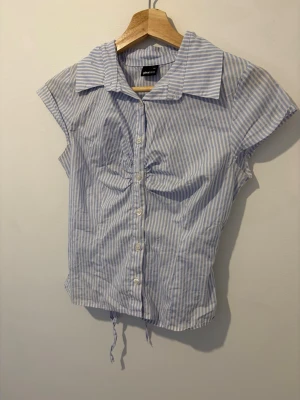 Blårandig kortärmad skjorta Gina Tricot - Snygg blårandig skjorta från Gina Tricot med korta ärmar och klassisk krage. Skjortan har knappar framtill och snörning i midjan för en cool detalj. Perfekt till jeans eller kjol för en fräsch look.