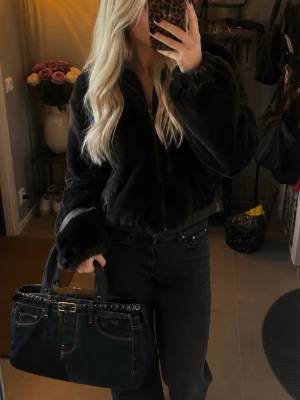 Svart denim handväska med bälte - Unik svart handväska i denim med detaljer som liknar ett par jeans, inklusive fickor och kontrastsömmar. Väskan har ett dekorativt bälte med spänne och svarta knappar längs ovankanten. Två rejäla handtag gör den enkel att bära över axeln.