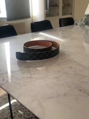 Svart och grått rutigt bälte - Snyggt bälte i svart och grått med rutigt mönster. Bältet har en klassisk design med svart spänne och insida i beige. Perfekt för att lyfta din outfit med en stilren accessoar.