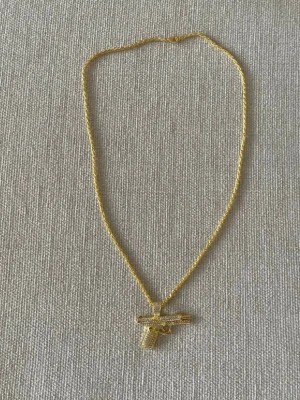 Guldhalsband med pistolhänge - Snyggt halsband i guldfärg med ett detaljerat pistolformat hänge täckt av glittrande stenar. Kedjan har en tvinnad design som ger extra bling och hänget är riktigt iögonfallande. Perfekt för dig som vill sticka ut med accessoarerna.