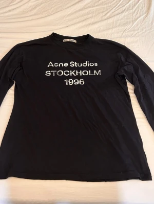 Svart långärmad t-shirt Acne Studios - Svart långärmad t-shirt från Acne Studios med tryck 'Acne Studios STOCKHOLM 1996' i vitt framtill. Klassisk rund hals och rak passform, perfekt för en clean och stilren look.