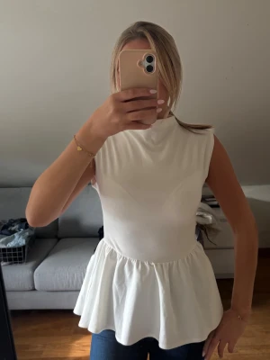 Vit peplum topp från Bik Bok - Säljer en vit peplum topp från Bik Bok med hög hals och ärmlös design. Toppen har en markerad midja och volang nedtill som ger en snygg siluett. Perfekt att styla med jeans eller kjol för en trendig look.