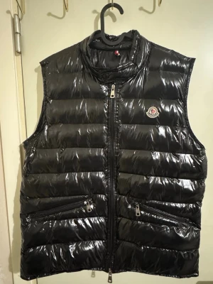 Svart quiltad dunväst från Moncler - Snygg svart dunväst från Moncler med glansig finish och quiltad design. Västen har hög krage, två dragkedjefickor och Moncler-logga på bröstet. Perfekt för lager-på-lager och håller dig varm under kyliga dagar.