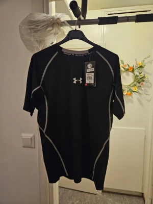 Svart träningströja från Under Armour - Snygg svart träningströja från Under Armour med korta ärmar och vita kontrastsömmar. Tillverkad i ett stretchigt funktionsmaterial som andas, perfekt för gymmet eller löprundan. Logga på bröstet och rund halsringning.