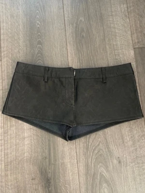 Svarta skinnshorts med bälteshällor - Säljer ett par svarta, tighta shorts i skinnimitation med bälteshällor och dragkedja fram. De har en lågmidjad passform och är perfekta för dig som vill ha en edgy och trendig look. Passar till många olika stilar och är riktigt statement.