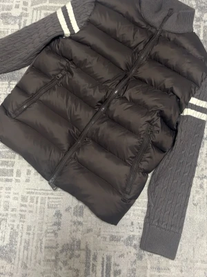 Moncler dunväst med stickade ärmar - Snygg dunväst från Moncler i mörkgrått med stickade, kabelmönstrade ärmar och vita ränder. Hög krage, två dragkedjefickor och ikonisk Moncler-logga på ärmen. Perfekt för lager-på-lager och streetstyle.