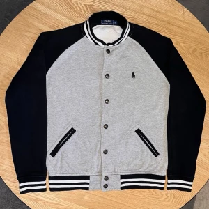 Polo Ralph Lauren Varsity Jacket Grå/Svart - Storlek M - Säljer en snygg Polo Ralph Lauren varsity-jacka i storlek M.  Grå body med svarta ärmar, klassiska vita ränder i krage och muddar.  Snap-knappar framtill och den ikoniska polo-loggan på bröstet.  Skick: Mycket bra. Tvättad och redo att användas. Inga synliga fläckar eller defekter.  Perfekt för en grisch/casual look.  Mått kan tas vid förfrågan.
