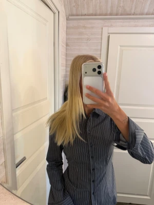 Mörkblå randig skjorta med knappar - Säljer en mörkgrå skjorta med vita tunna ränder och knappar framtill. Skjortan har lång ärm och klassisk krage, samt markerad midja för en snygg passform. Perfekt till jeans eller kjol för en clean look.
