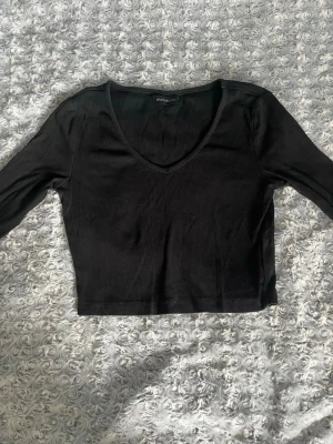 Svart croppad långärmad topp - Snygg svart croppad topp med lång ärm och v-ringning. Tillverkad i ett ribbat material som ger en skön och stretchig passform. Perfekt att styla med höga jeans eller kjol för en trendig look.