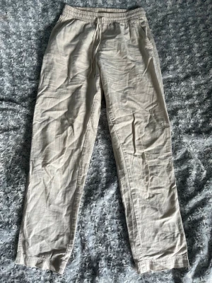 Beiga linnebyxor med resårmidja - Säljer ett par ljusbeige linnebyxor med avslappnad passform och resår i midjan samt dragsko. Byxorna har raka ben och sidofickor, perfekta för en chill och luftig stil. Materialet är linne som ger en skön känsla under varma dagar.
