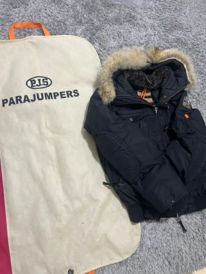 Svart dunjacka från Parajumpers - Snygg svart dunjacka från Parajumpers med avtagbar pälskant på huvan och flera praktiska fickor. Jackan har quiltat foder, orange detaljer och patch på ärmen. Perfekt för kalla vinterdagar och riktigt varm.