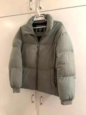 Grå pufferjacka från FSBN XS - Säljer en stilren grå pufferjacka från FSBN i storlek XS. Jackan har hög krage, dragkedja framtill och två sidofickor. Perfekt för kalla vinterdagar och ger en clean look till din outfit. Materialet är syntetiskt och jackan har ribbade muddar vid ärmsluten.