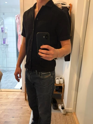 Svart kortärmad skjort-tshirt - Säljer en svart kortärmad t-shirt med skjortkrage och knappar framtill. Materialet är strukturerat och ger en unik känsla. Perfekt till jeans eller shorts för en avslappnad men stilren look.