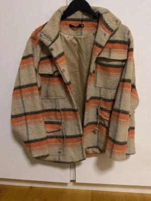 Beige overshirt med orange rutor - Snygg overshirt i beige med orange och mörkgröna rutiga ränder. Jackan har fyra fickor framtill, dragsko i midjan och en mjuk, ullig yta. Perfekt för lager på lager och ger en avslappnad men trendig look.