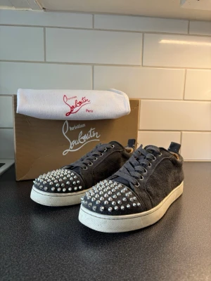 Svarta sneakers med nitar från Louboutin - Snygga svarta sneakers i mocka från Christian Louboutin med silverfärgade nitar på tån och vit sula. Skorna har klassisk låg profil, svarta skosnören och en unik edgy look som sticker ut. Perfekta för dig som vill ha något extra till din outfit. Har haft defekt vid insidan sulan mot hälen men fixade det hos en skomakare som lade in en läder lapp syns på bild 3. Vet ej om äkhekt därav priset!