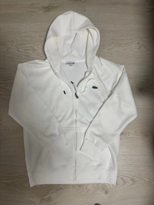 Vit hoodie med dragkedja från Lacoste - Snygg vit hoodie från Lacoste med dragkedja framtill och klassisk krokodillogga på bröstet. Hoodien har huva med dragsko och två fickor framtill. Perfekt för en clean och stilren look.