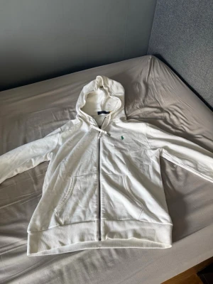 Vit hoodie med dragkedja från Polo Ralph Lauren - Snygg vit hoodie från Polo Ralph Lauren med dragkedja framtill och klassisk grön logga broderad på bröstet. Hoodien har huva med snören och två fickor framtill. Tillverkad i mjukt bomullsmaterial som är skönt att ha på sig.