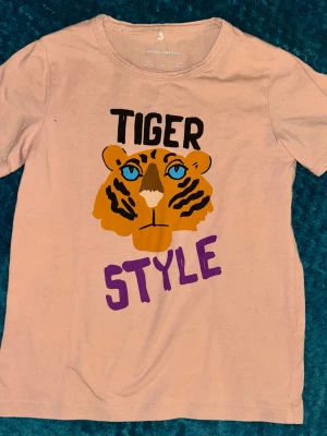 Rosa t-shirt med tigertryck Mini Rodini - Rosa t-shirt från Mini Rodini med tryck av en tiger och texten 'Tiger Style' i svart och lila. T-shirten har rund hals och korta ärmar. Perfekt för dig som gillar coola och lekfulla prints.