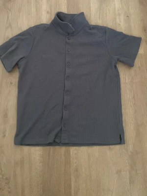 Grå kortärmad skjorta med hög krage - Säljer en stilren grå skjorta med korta ärmar och hög krage. Skjortan har knappar hela vägen framtill och är tillverkad i ett mjukt material som känns bekvämt mot huden. Perfekt för dig som vill ha en enkel men unik look.