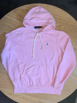 Polo Ralph Lauren Hoodie Rosa – Unisex – Fint skick - Säljer en riktigt fin Polo Ralph Lauren hoodie i ljusrosa.  Klassisk modell med liten blå polo-logo på bröstet, huva med vita snören och känguruficka.  Skick: Mycket bra. Tvättad och klar. Inga synliga defekter. Bra kvalitet.  Unisex – passar både killar och tjejer.  Storlek: M (något mindre i storleken) Mått kan tas vid förfrågan.