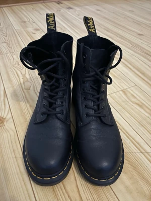 Svarta Dr. Martens läderstövletter - Klassiska svarta stövletter från Dr. Martens i äkta läder med snörning framtill och ikoniska gula sömmar runt sulan. Grov yttersula och dragflikar med logga baktill ger en cool och tidlös look.