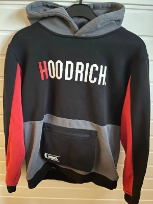 Svart och röd hoodie från Hoodrich - Snygg hoodie från Hoodrich i svart, grått och rött med stor logga på bröstet. Hoodien har huva, magficka med dragkedja och röda detaljer längs ärmarna. Perfekt för dig som gillar streetwear och vill sticka ut med cool design. Det är xl u barn storlek, så det motsvarar S i vuxen