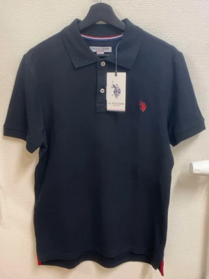 Svart pikétröja från U.S. Polo Assn - Klassisk svart pikétröja från U.S. Polo Assn med korta ärmar och krage. Tröjan har två knappar framtill och en röd broderad logga på bröstet. Tillverkad i bomull för en bekväm känsla och stilren look.