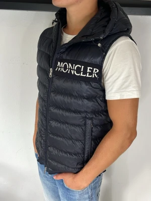 Moncler väst - Snygg svart dunväst från Moncler med stor vit logga på bröstet. Västen har en vadderad design, dragkedja framtill, två fickor med dragkedja och en rejäl huva. Perfekt för lager-på-lager och streetstyle.