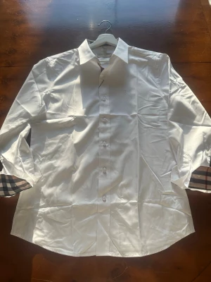 Vit burberry skjorta  - Klassisk vit skjorta i bomull med långa ärmar och knappar framtill. Manschetterna har en snygg rutig detalj i beige, svart och rött som ger en unik touch. Perfekt för dig som vill ha en clean look med lite extra stil.