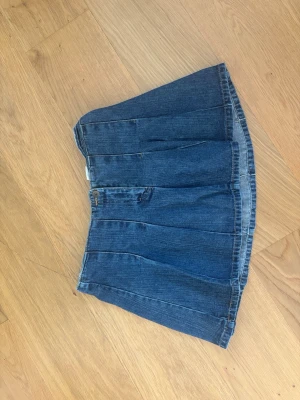 Kort jeanskjol i blå denim - Säljer en kort jeanskjol i klassisk blå denim med veckad design. Kjolen har knapp och dragkedja framtill och är perfekt för dig som gillar en avslappnad och trendig stil. 