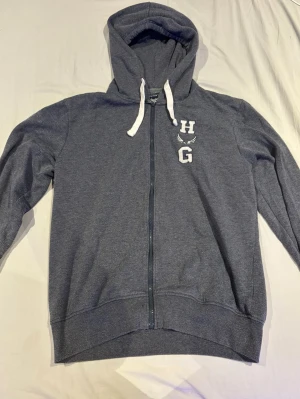 Mörkgrå zip-hoodie med vita snören - Mörkgrå hoodie med dragkedja och huva, vita snören och tryck med bokstäverna H och G på bröstet. Mjuk och skön bomullsblandning, perfekt för en avslappnad stil. Långa ärmar och ribbade muddar vid ärmslut och nederkant.