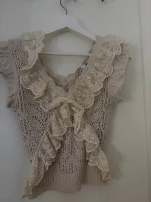 Beige stickad topp med spetsvolang - Supersöt beige stickad topp med v-ringning och stora spetsvolanger framtill och runt halsen. Toppen är ärmlös och har ett romantiskt, vintageinspirerat utseende med fina virkade detaljer och en dekorativ rosett framtill.