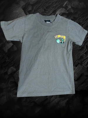 Stussy tröja  - Säljer en grå t-shirt från Stüssy med coolt tryck av tärningar och gul logga både fram och bak. T-shirten har normal passform och är kortärmad, perfekt för dig som gillar streetwear och vill sticka ut lite extra. Använd men i bra skick 